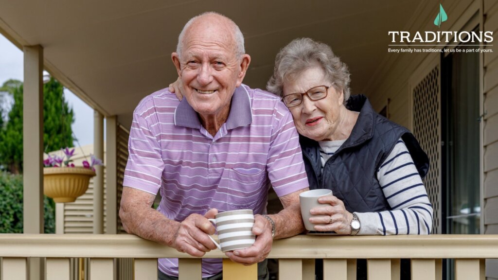 An elderly couple standing outside their house holding coffee cups with the traditions logo on the top right corner