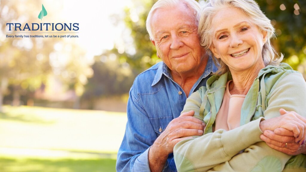 An elderly couple hugging each other outside with the Traditions logo at the top left corner.