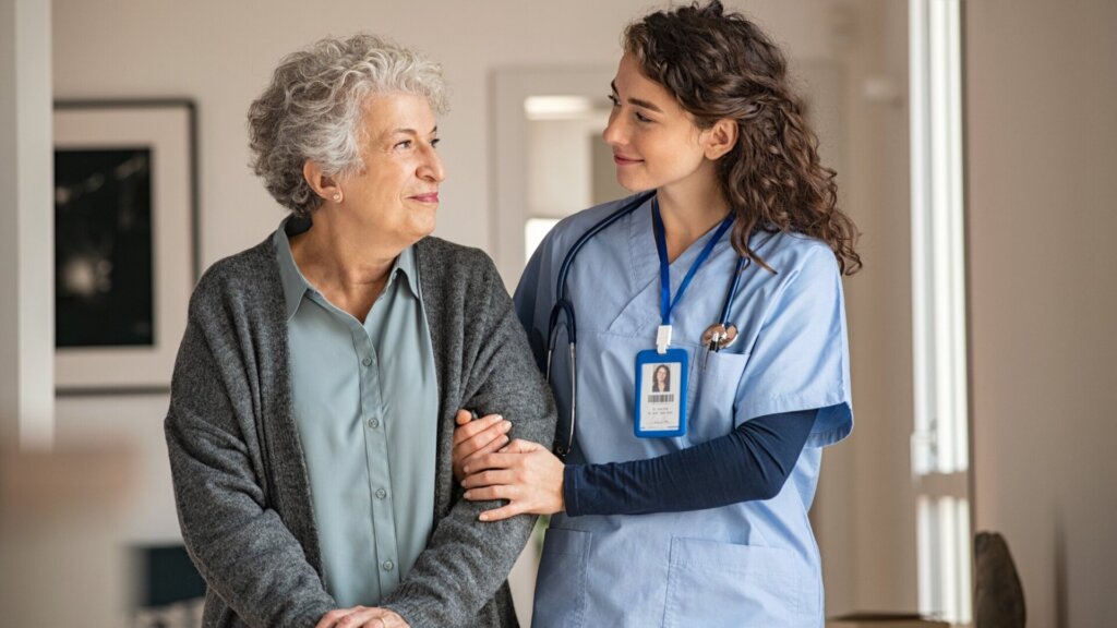A woman and caregiver walking together down a hallway holding and looking at each other.
