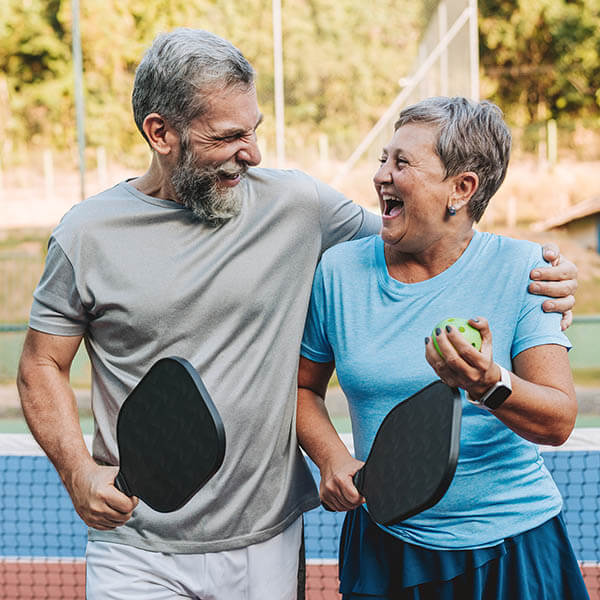 Senior man and senior woman playing pickleball and laughing outdoors at Mission Grove at Stoneybrook senior living community