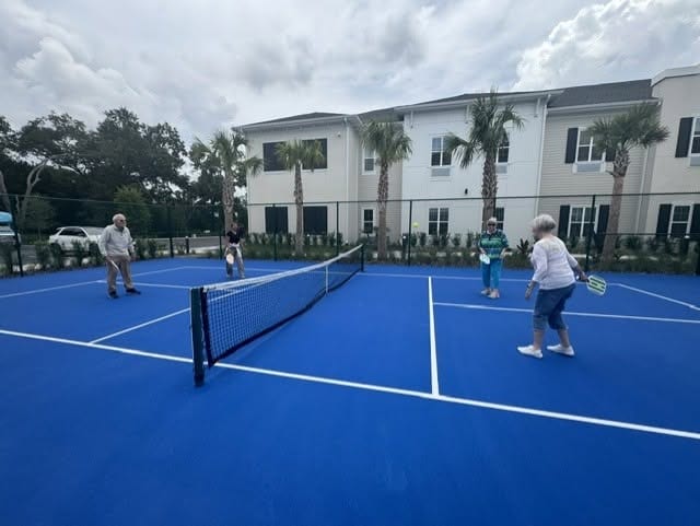 Four residents playing doubles pickleball on a blue outdoor court at Mission Grove at Stoneybrook, with palm trees and the community building visible in the background.