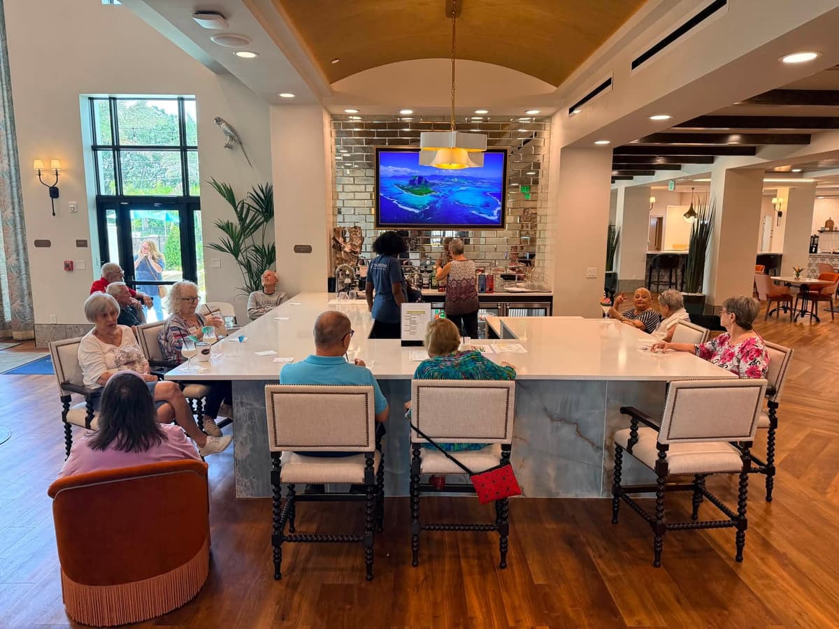 Residents gather around a large bar island in Mission Grove at Stoneybrook's elegantly appointed clubroom during a piña colada social event, with staff serving drinks nearby.