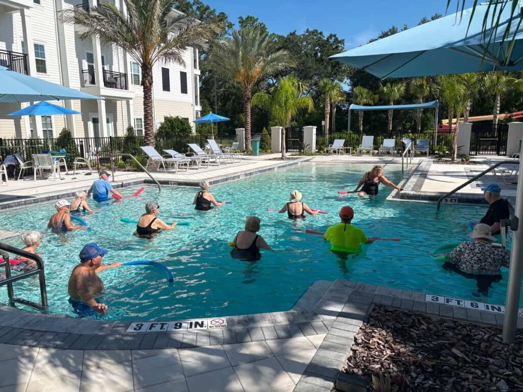A group of residents participates in an outdoor water aerobics using foam noodles in the resort-style pool at Mission Grove at Stoneybrook, surrounded by palm trees and lounge chairs.